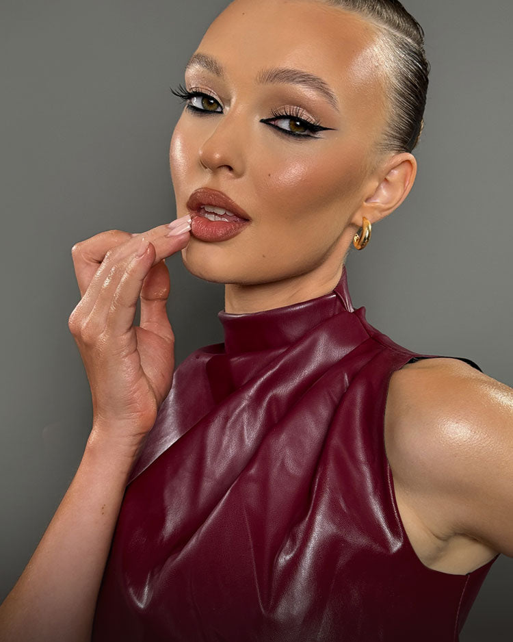 Beautiful female model with bronzed makeup, winged eyeliner and brown lipstick wearing a burgundy leather top.