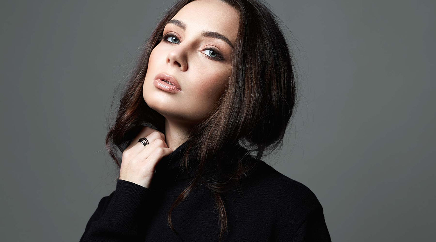 Woman with dark hair, soft matte makeup, wearing a black top against a grey background.