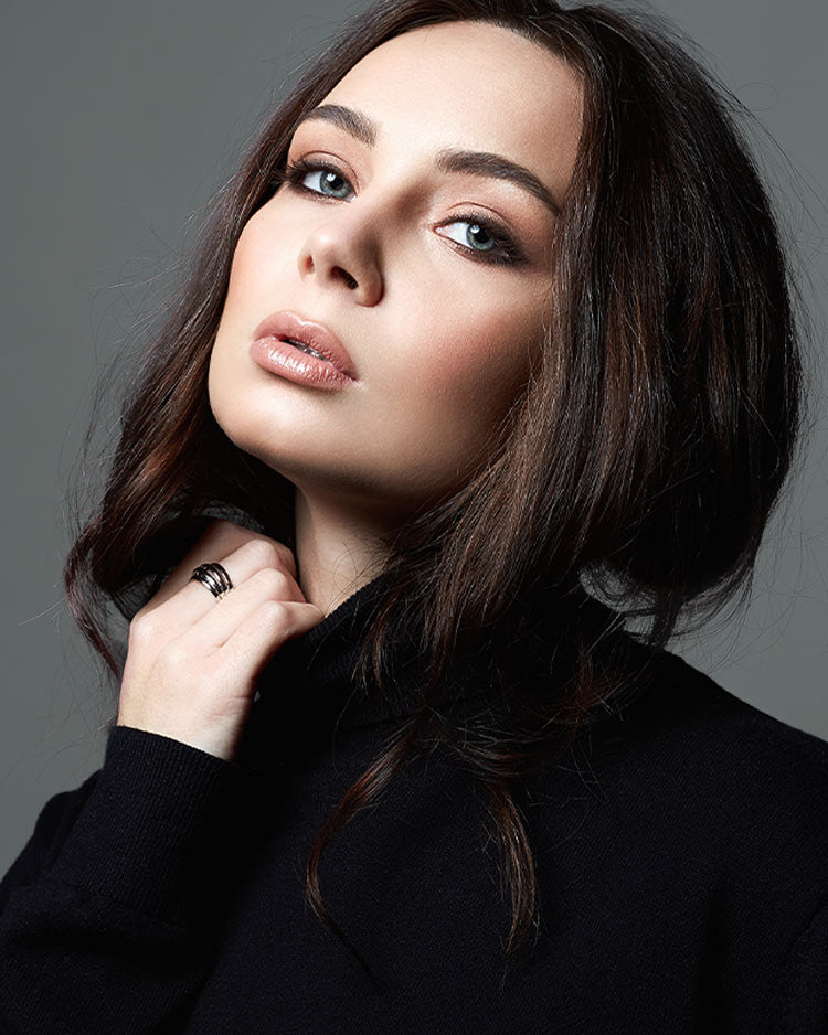 Woman with dark hair, soft matte makeup, wearing a black top against a grey background.