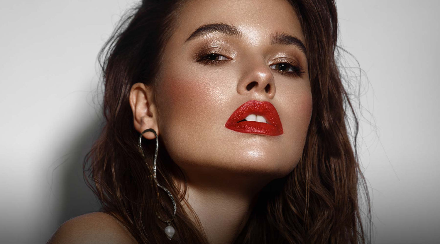 Dark haired female model with bronze, glowing makeup, bright red lipstick and wearing large statement earrings.
