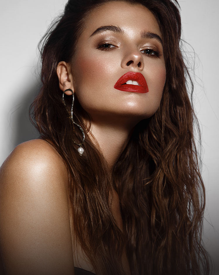 Dark haired female model with bronze, glowing makeup, bright red lipstick and wearing large statement earrings.
