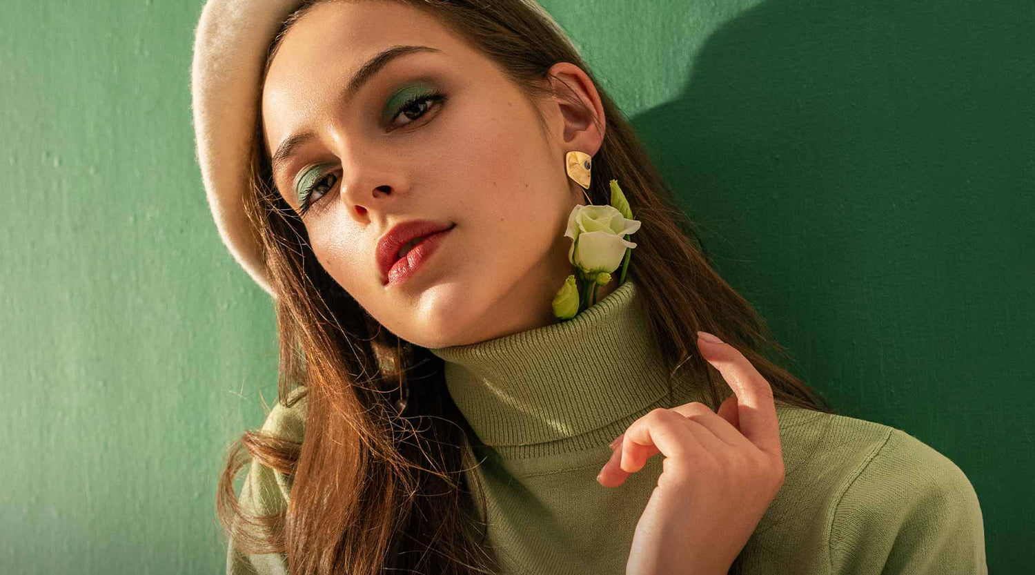 Female with green eyeshadow, brown lipstick wearing a green jumper and floral earrings against a green background.