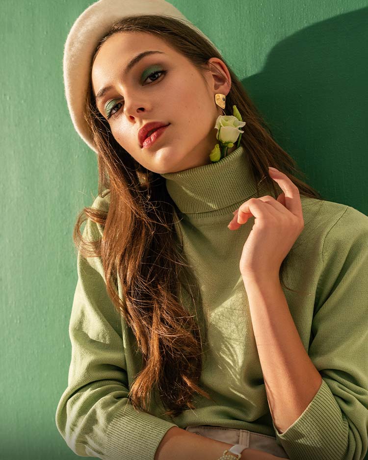 Female with green eyeshadow, brown lipstick wearing a green jumper and floral earrings against a green background.