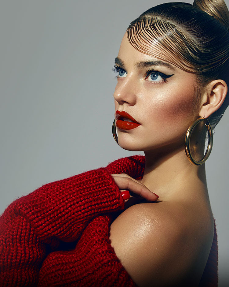 Beautiful female model with bronzed glam makeup, winged eyeliner, red lipstick with sleek styled hair and wearing red jumper.