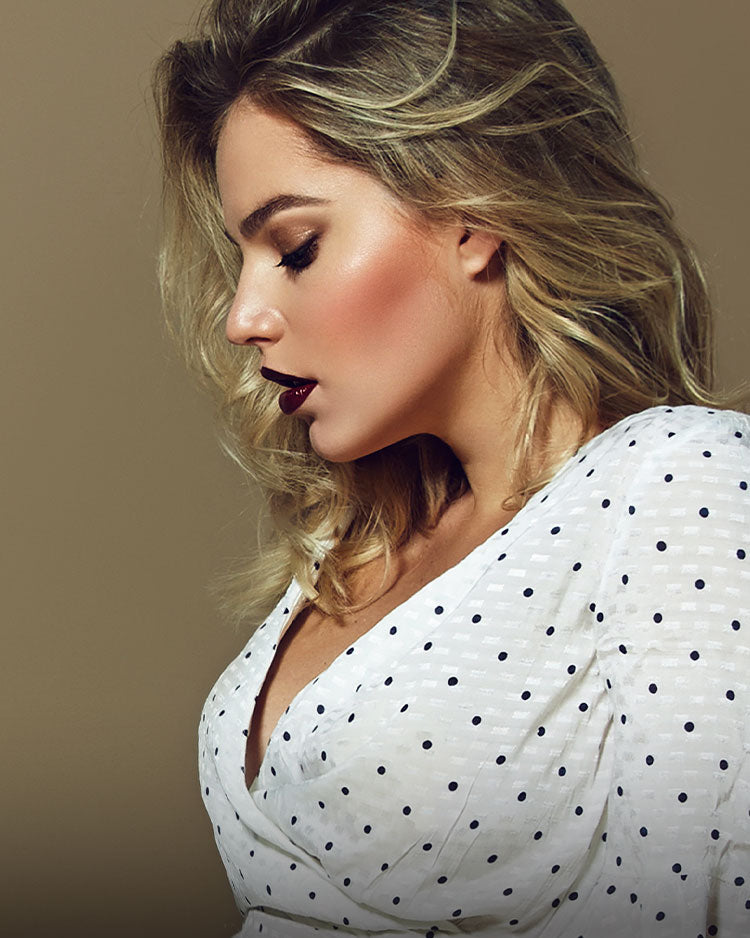 Beautiful blonde model with gold eyeshadow, blushed cheeks, dark plum lipstick wearing a polka dot blouse.