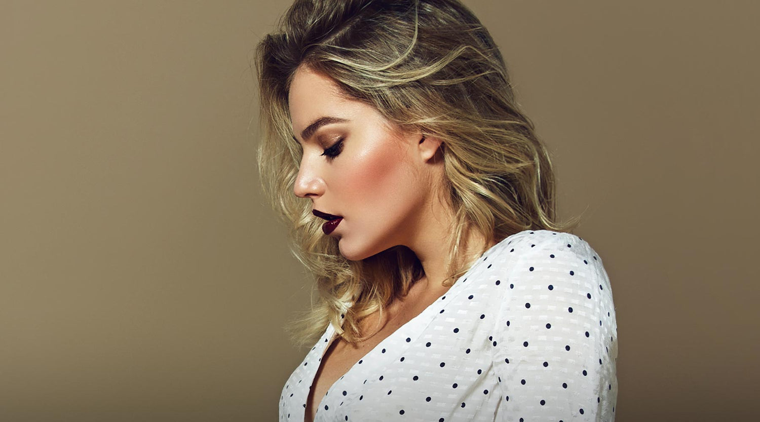 Beautiful blonde model with gold eyeshadow, blushed cheeks, dark plum lipstick wearing a polka dot blouse.