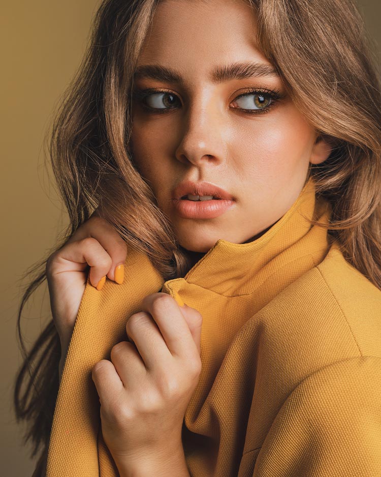 Beautiful female with gold eyeshadow and nude lipstick, wearing a yellow jacket on a soft green background.