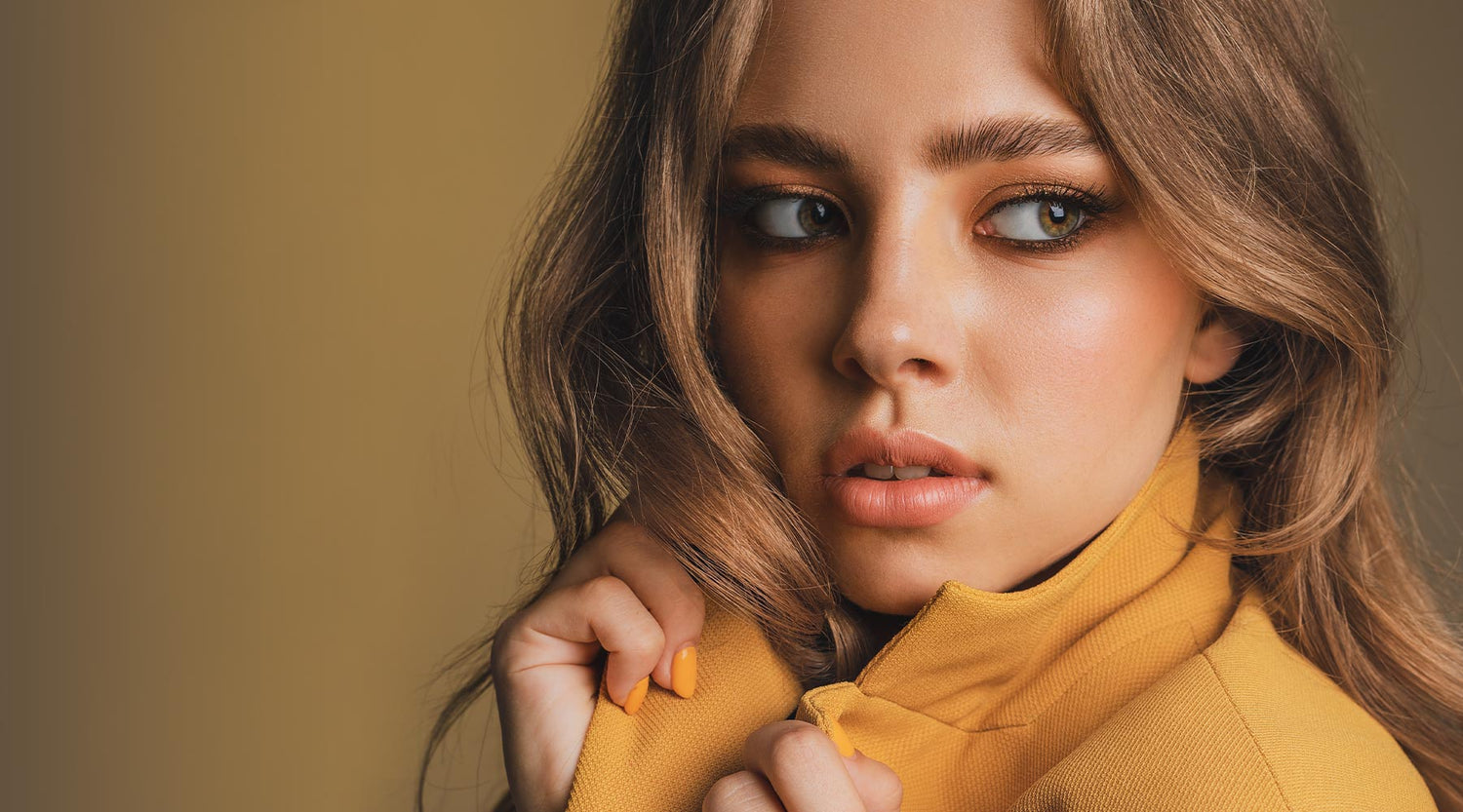Beautiful female with gold eyeshadow and nude lipstick, wearing a yellow jacket on a soft green background.